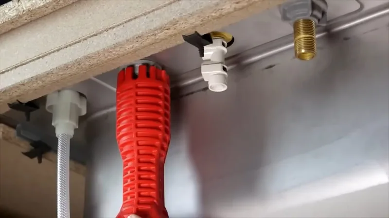 Red basin wrench held up to white and brass fittings beneath a sink, with a braided supply line at the left