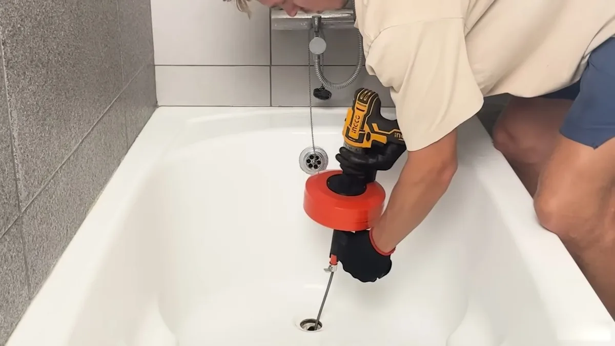 Technician in work gloves feeding a drill-driven drum auger into a white bathtub drain
