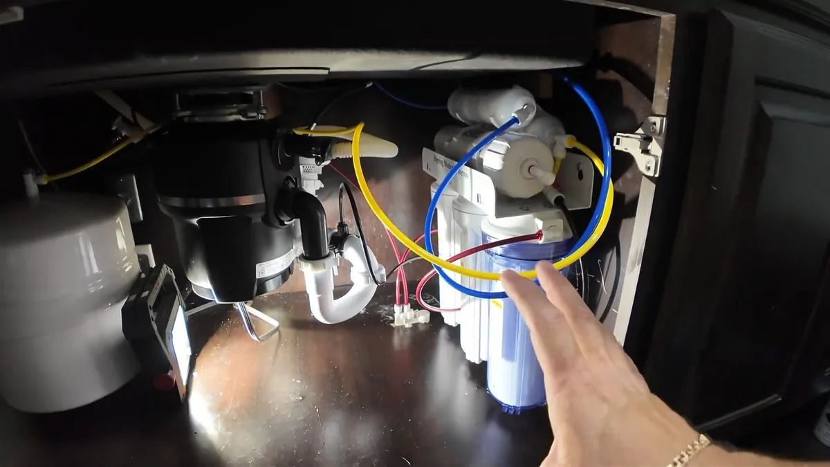 Open under-sink cabinet with a black garbage disposal, a white drain trap and a three-stage water filter with blue and yellow tubing