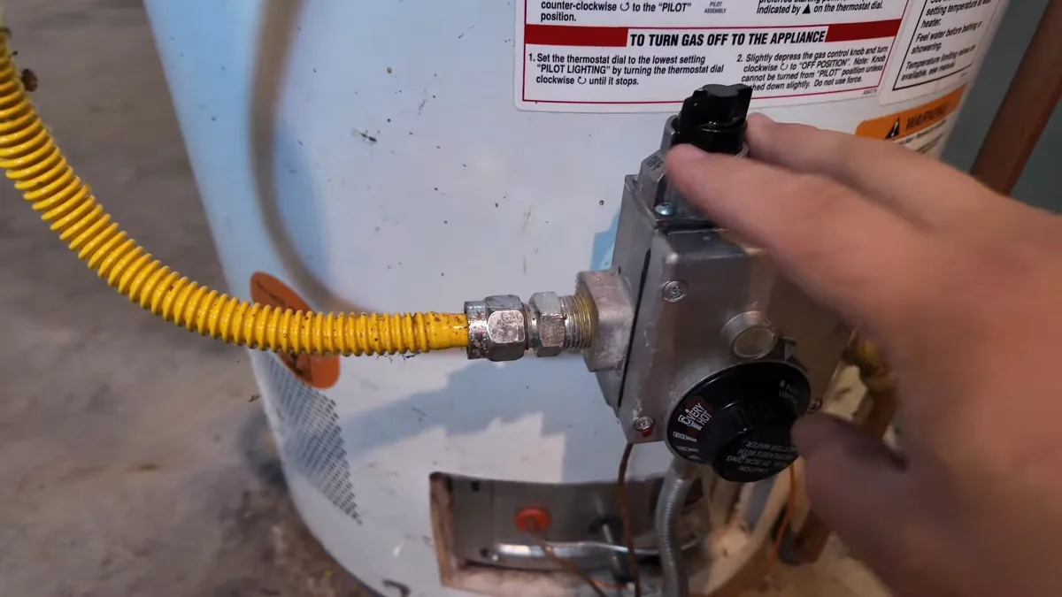 Hand on the black knob of a gas control valve on a tank water heater, with a yellow flexible gas line entering from the left
