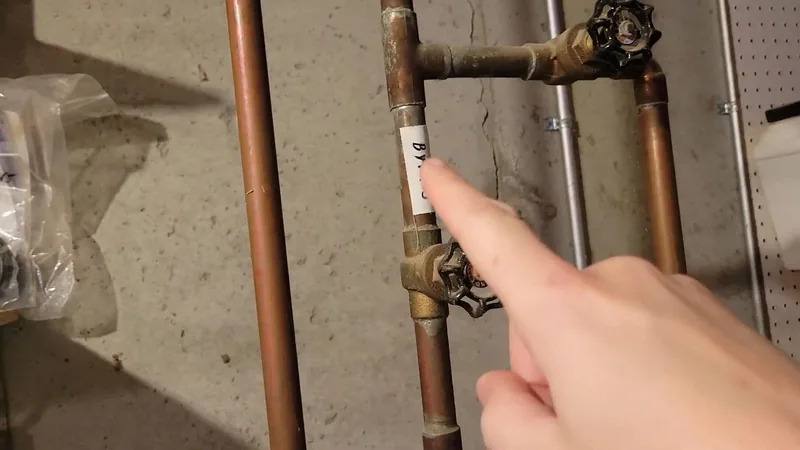 Hand pointing at a brass valve on a copper supply pipe in a basement, with a second valve on a branch above it