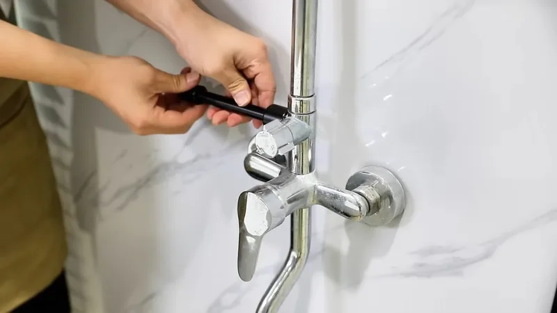 Hands holding a small black tool against a chrome wall-mounted shower mixer valve on a marble-look wall