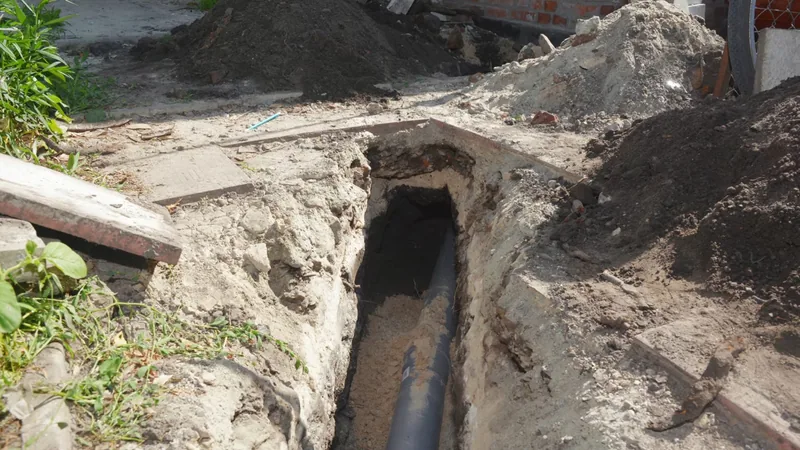 Narrow trench in a yard with a gray pipe running along its bottom and piles of excavated soil on both sides