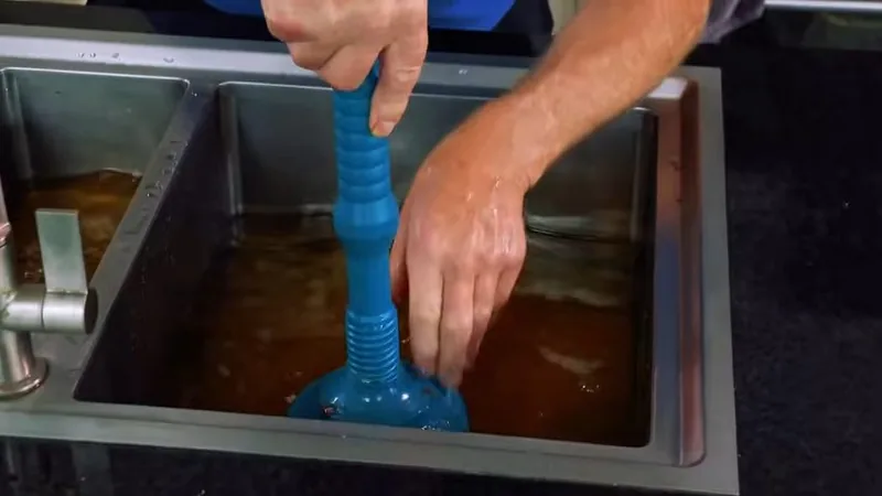 Hands working a blue plunger in a stainless double sink filled with murky standing water