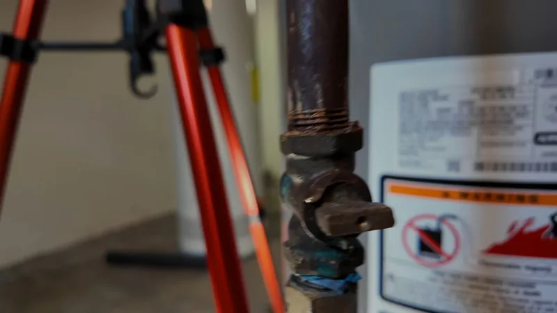 Rating plate and warning label on the side of a gas tank water heater, beside a corroded gas shutoff valve and a red tripod