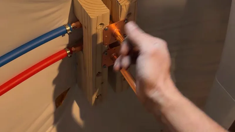 Red and blue PEX lines clamped to copper stub-outs on a wood mounting block, with a hand steadying one stub