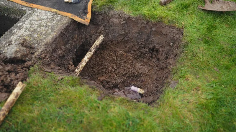 Small excavated pit in a lawn with a shovel nearby, wooden stakes lying across the soil and a pale pipe end at the bottom