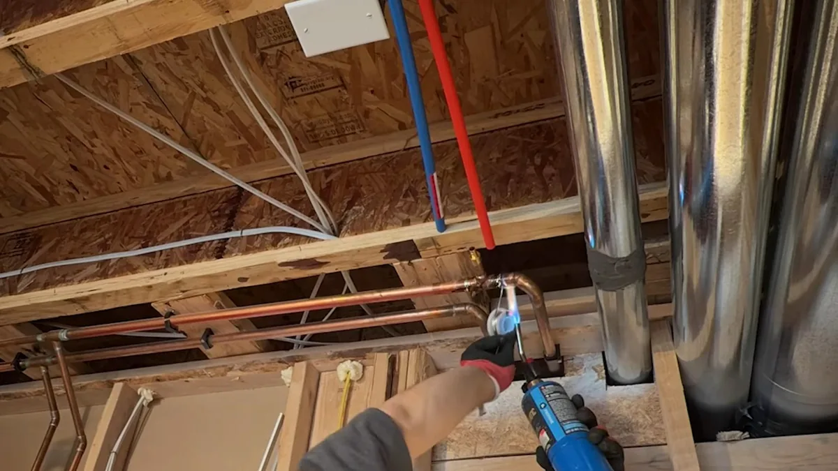 Technician in a glove using a torch on a copper fitting between open joists, with red and blue PEX lines running overhead