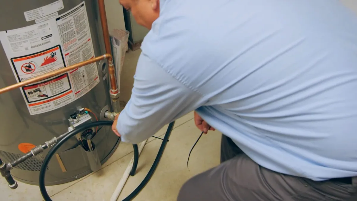 Technician in a light blue shirt crouching at the lower connections of a gas tank water heater with a black hose on the floor