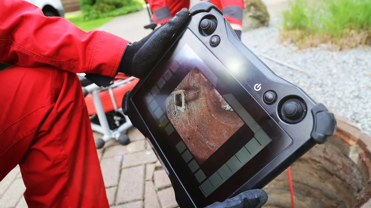Technician in red coveralls holding a drain camera monitor that shows the inside of a pipe, beside an open yard access