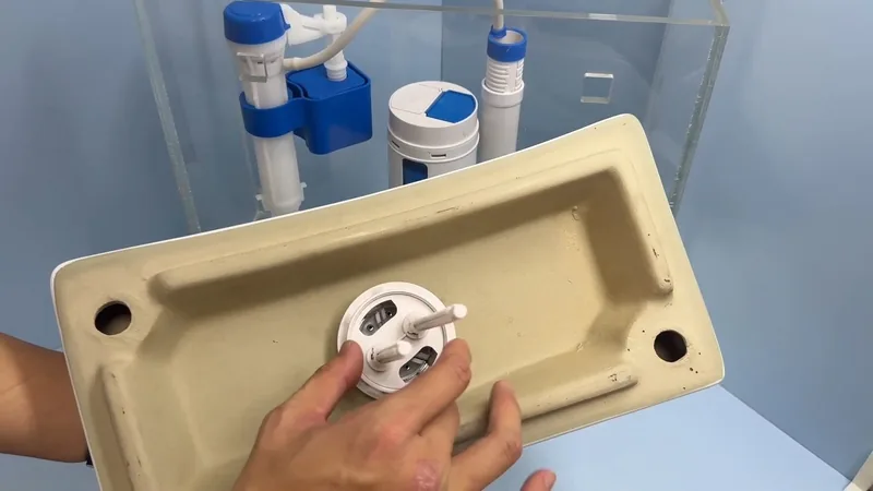 Hands holding a ceramic tank lid upside down beside a clear tank model with a fill valve and other parts inside