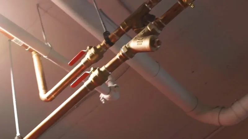 Two red-handled brass ball valves on copper supply lines running along a ceiling beside a white drain pipe