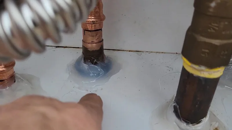 Top of a white tank water heater with a copper stub, a braided flex connector and a brown iron pipe wrapped in yellow tape