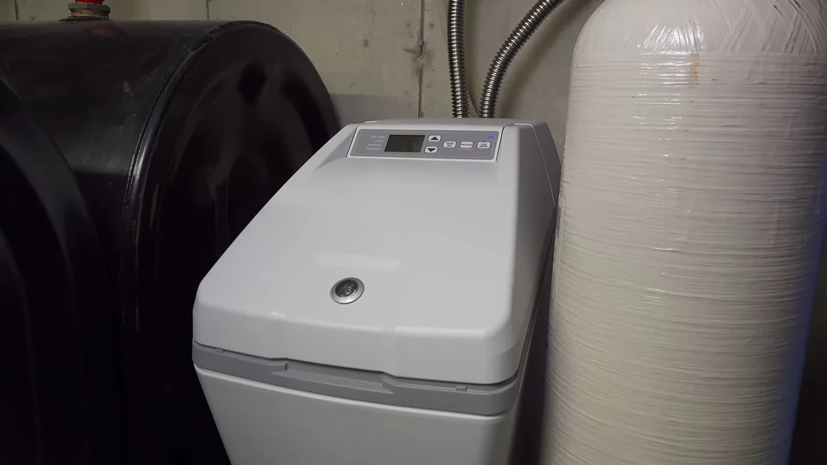 Gray water softener with a digital control panel beside a tall white resin tank and a black tank against a concrete wall